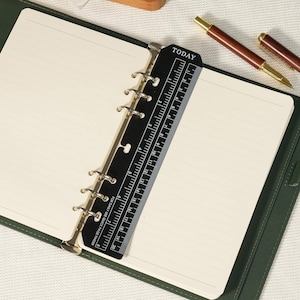 May include: A dark green ring-bound planner with cream-colored lined paper and a black plastic page marker. The marker features the word TODAY at the top, a ruler with markings up to 8 inches and 20 cm, and the text FOR DAILY USE QUICK PAGE FINDER.