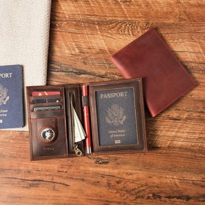 May include: A dark brown leather passport holder featuring multiple card slots, a zippered pocket, a pen loop with a red pen, and a circular tracking device slot. A United States passport is inserted into the clear window. A separate matching leather cover lies nearby.
