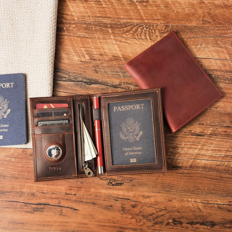 May include: A dark brown leather passport holder featuring multiple card slots, a zippered pocket, a pen loop with a red pen, and a circular tracking device slot. A United States passport is inserted into the clear window. A separate matching leather cover lies nearby.