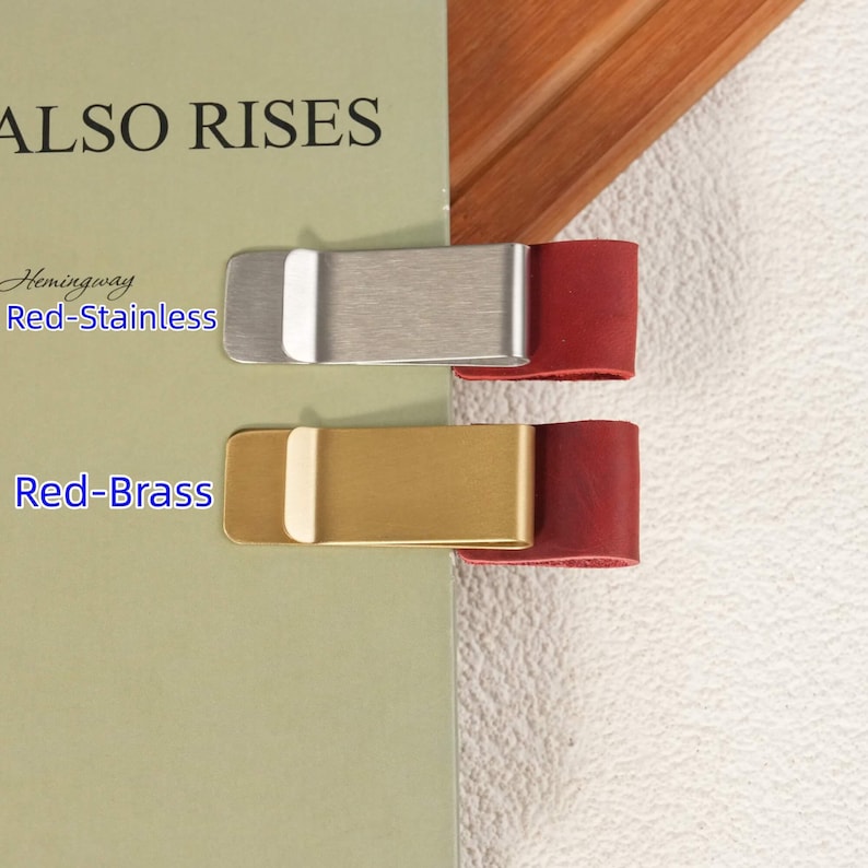 May include: Two metal money clips attached to a book page. The top clip is brushed stainless steel with a red leather tab, labeled Red-Stainless. The bottom clip is gold-toned brass with a red leather tab, labeled Red-Brass. The book cover text reads ALSO RISES.