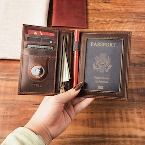 May include: A brown leather passport holder featuring multiple card slots, a zippered pocket, a pen loop with a red pen, and a dedicated circular compartment for a tracking device. The right side displays a United States passport with the text PASSPORT.