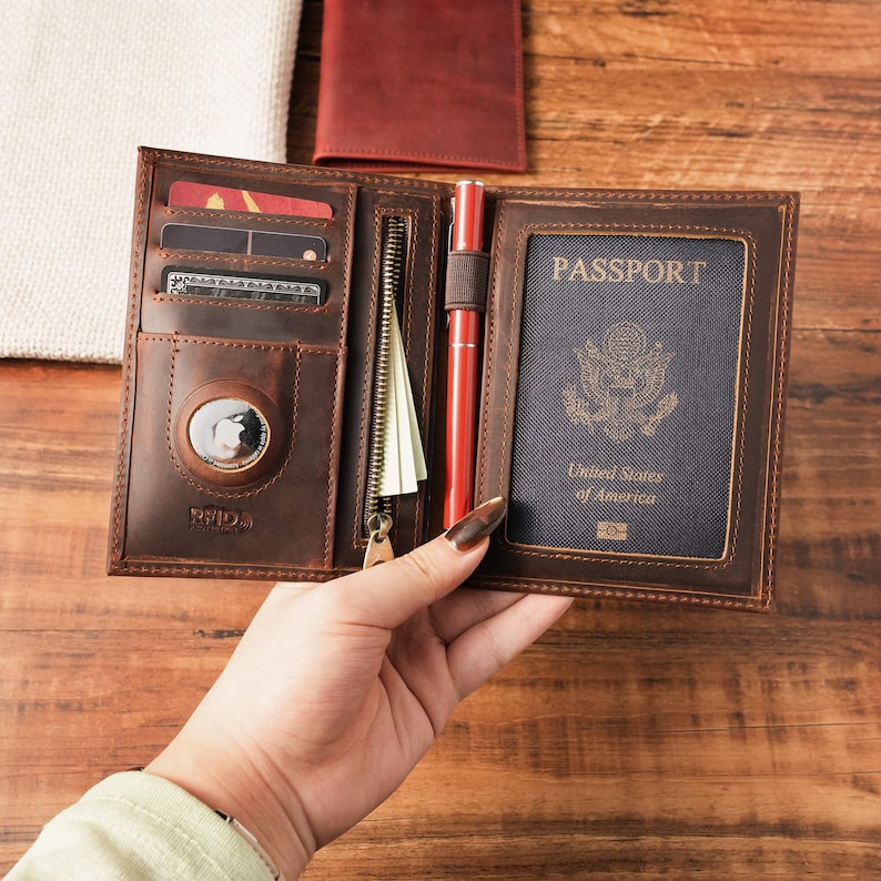 May include: A brown leather passport holder featuring multiple card slots, a zippered pocket, a pen loop with a red pen, and a dedicated circular compartment for a tracking device. The right side displays a United States passport with the text PASSPORT.