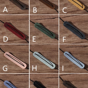 May include: Twelve leather cord keepers in various colors, including brown, black, red, green, pink, blue, and gray. The cord keepers are arranged in a grid pattern on a wooden surface.
