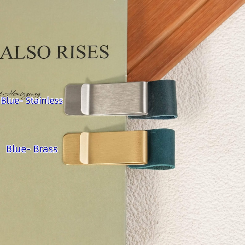 May include: Two metal money clips attached to a book cover. The top clip is brushed stainless steel with a dark blue leather loop, and the bottom clip is brushed brass with a dark blue leather loop. Text on the book reads ALSO RISES.