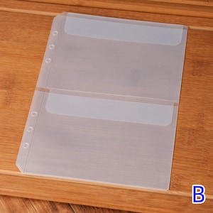 May include: Clear plastic binder sheet with two pockets. The sheet has three holes on the left side for insertion into a binder. The pockets have a frosted top edge. The sheet is on a wooden surface.