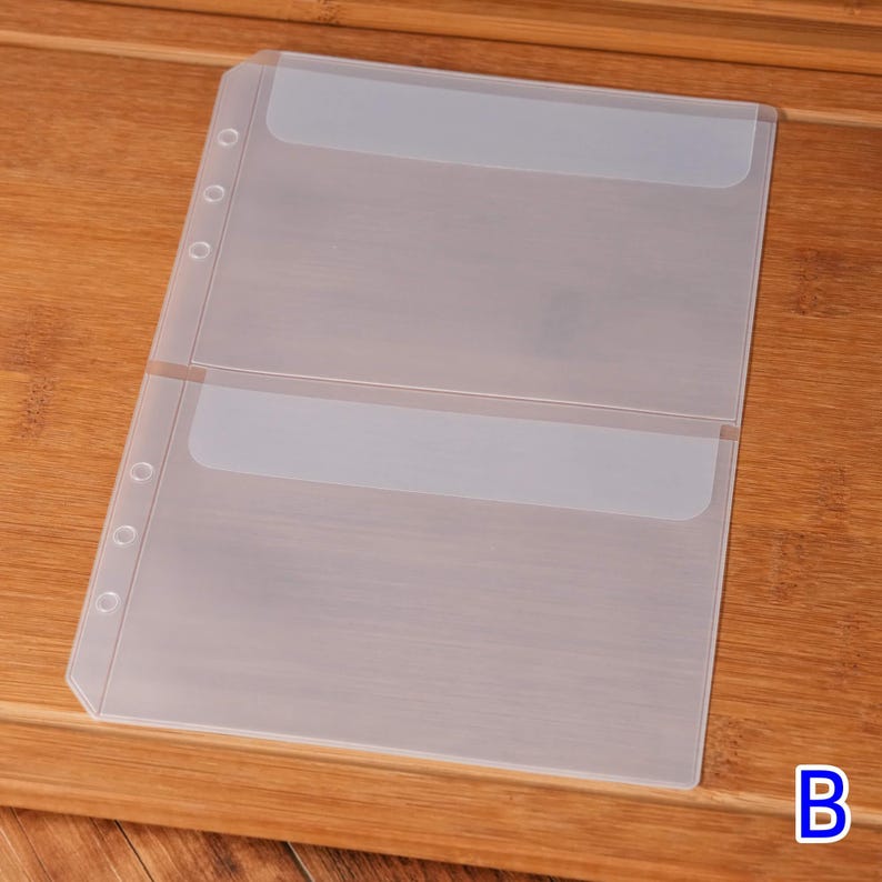 May include: Clear plastic binder sheet with two pockets. The sheet has three holes on the left side for insertion into a binder. The pockets have a frosted top edge. The sheet is on a wooden surface.