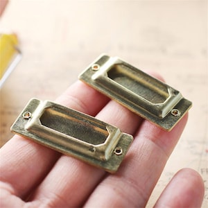 May include: Two gold-toned metal label holders with a rectangular shape and a slot for inserting a label. The holders have a smooth, polished finish.
