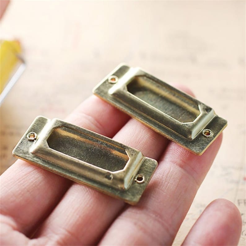 May include: Two gold-toned metal label holders with a rectangular shape and a slot for inserting a label. The holders have a smooth, polished finish.