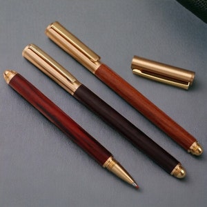 Könnte beinhalten: Drei Holzstift mit goldenen Akzenten. Die Stifte haben eine dunkle Holz- und eine helle Holzoptik. Die Stifte befinden sich auf einer grauen Oberfläche.