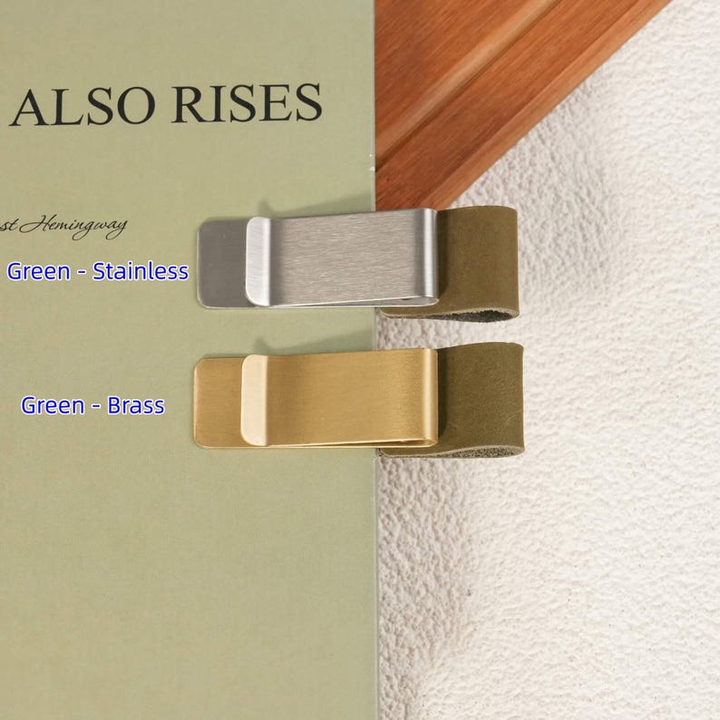 May include: Two rectangular metal money clips attached to a green book cover. The top clip is brushed stainless steel, and the bottom clip is brass. Each clip features a dark green leather tab extending from the end, providing a secure grip.