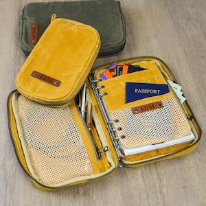 May include: Mustard yellow travel organizer with a zippered closure, mesh pockets, and a leather tag that reads "HIM." The organizer is open, revealing a passport, cards, and pens. A smaller matching pouch and a green pouch are also visible.