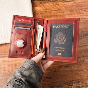 May include: A red leather passport holder featuring multiple card slots, a dedicated pocket for a tracker device, a zippered compartment containing paper currency, and a pen loop holding a red pen. A United States passport is inserted on the right.
