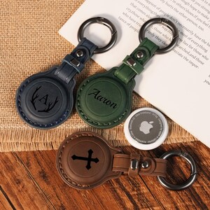 May include: Three circular leather keychains with metal rings, featuring a dark blue one with an antler and letter A design, a green one with the name Aaron, and a brown one with a cross design, alongside a white circular tracking device.