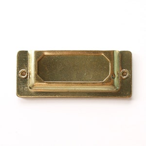 May include: A gold metal label holder with a rectangular shape and rounded corners. The holder has two small holes on each side for attaching it to a surface.