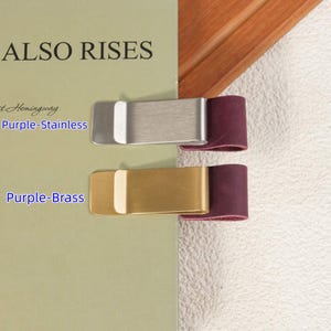 May include: Two metal money clips attached to a book cover. The top clip is silver-toned stainless steel with a purple leather loop, and the bottom clip is gold-toned brass with a purple leather loop. Text reads Purple-Stainless and Purple-Brass.
