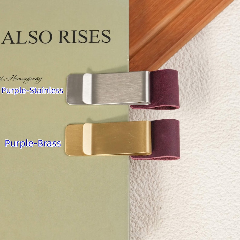 May include: Two metal money clips attached to a book cover. The top clip is silver-toned stainless steel with a purple leather loop, and the bottom clip is gold-toned brass with a purple leather loop. Text reads Purple-Stainless and Purple-Brass.