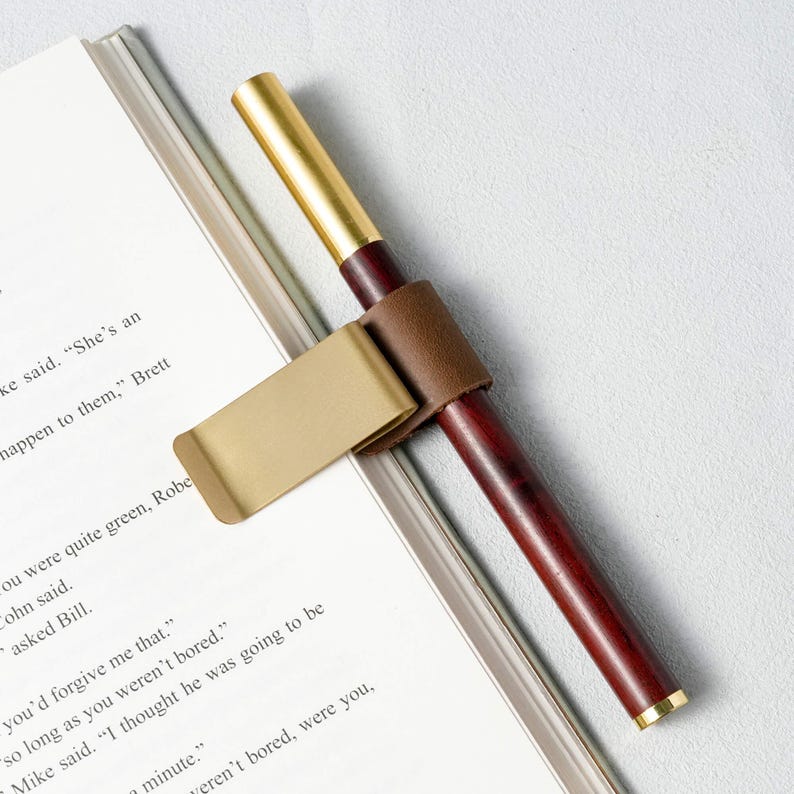 May include: A wooden pen with a gold-colored tip and a dark brown barrel, held in place by a brown leather pen holder attached to a gold-colored clip. The pen rests on a white page with printed text.