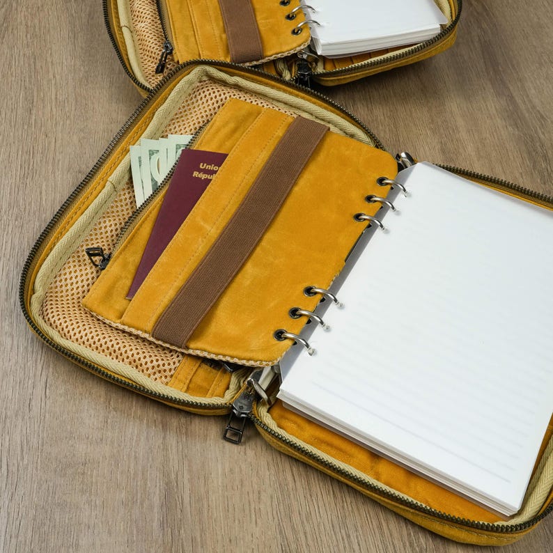 May include: Open mustard-yellow zippered organizer with a brown elastic strap and a matching notebook. Inside, a passport and cash are visible in a mesh pocket. The notebook has lined white pages. The organizer is on a wooden surface.