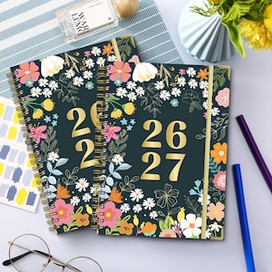 May include: Two spiral-bound planners with floral designs and the years 2026 and 2027 in gold. The covers are dark green with colourful flowers and gold accents. The planners are open on a light blue surface.