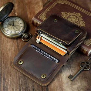 Op de afbeelding: Een bruine leren portemonnee met ritssluiting, open om kaarten en contant geld te laten zien. De portemonnee ligt op een houten oppervlak naast een vintage zakhorloge, een boek en een decoratieve sleutel.