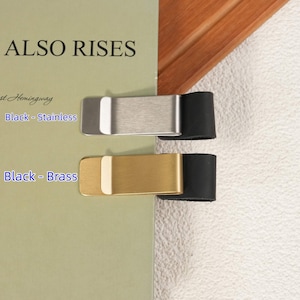 May include: Two money clips attached to a book cover. The top clip is silver-toned stainless steel with a black leather-like end. The bottom clip is gold-toned brass with a black leather-like end. Text on the book reads ALSO RISES, Black - Stainless, and Black - Brass.