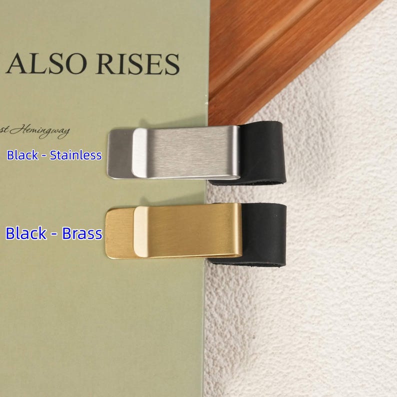 May include: Two money clips attached to a book cover. The top clip is silver-toned stainless steel with a black leather-like end. The bottom clip is gold-toned brass with a black leather-like end. Text on the book reads ALSO RISES, Black - Stainless, and Black - Brass.