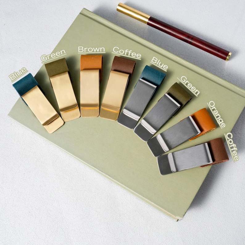 May include: A collection of metal money clips in various colors, including blue, green, brown, coffee, and orange, arranged on a light green book. A pen with a gold tip and a dark brown barrel rests on the book.