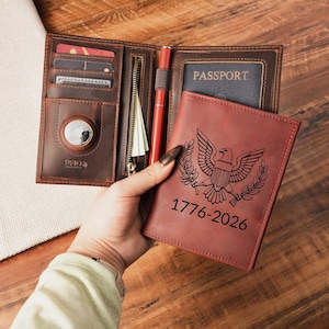 May include: A brown leather passport wallet featuring an interior AirTag slot, card pockets, a zippered compartment, and a pen loop. The exterior cover is engraved with an eagle emblem and the text 1776-2026, with a passport inserted inside.
