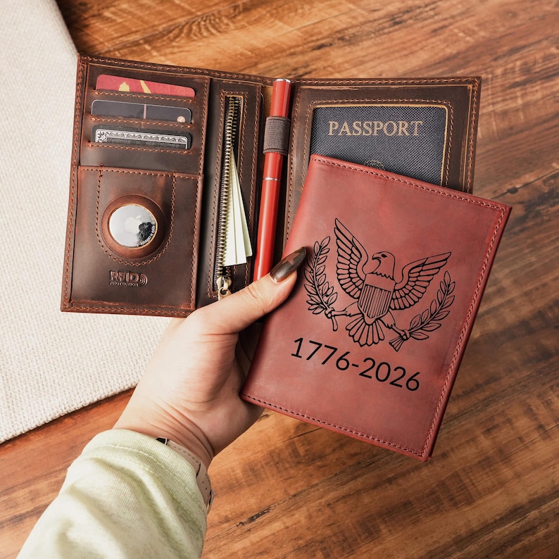 May include: A brown leather passport wallet featuring an interior AirTag slot, card pockets, a zippered compartment, and a pen loop. The exterior cover is engraved with an eagle emblem and the text 1776-2026, with a passport inserted inside.