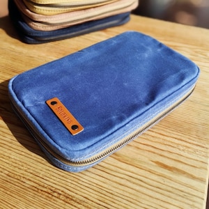 May include: A blue rectangular zippered pouch with a brown leather tag that reads "SOLOKING". The pouch is on a wooden surface, with other pouches visible in the background. The zipper is a light brown color.