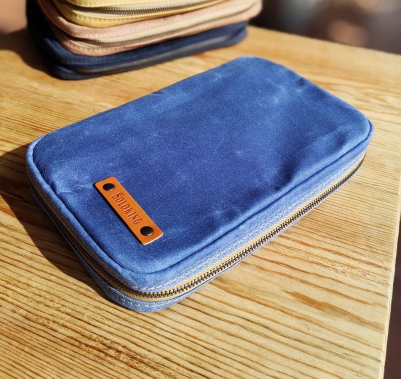 May include: A blue rectangular zippered pouch with a brown leather tag that reads "SOLOKING". The pouch is on a wooden surface, with other pouches visible in the background. The zipper is a light brown color.