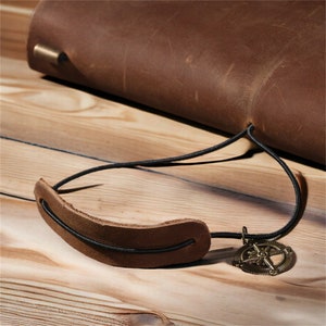 May include: A brown leather journal with a black elastic closure and a compass charm. The journal is on a wooden surface.