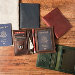 May include: A collection of leather passport holders in brown, dark blue, red, and green. The open brown holder features slots for credit cards, a zippered pocket, a pen loop with a red pen, and a dedicated circular compartment for a tracker.