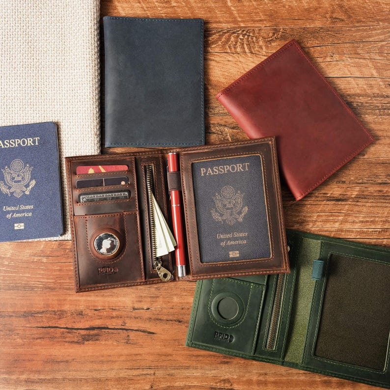 May include: A collection of leather passport holders in brown, dark blue, red, and green. The open brown holder features slots for credit cards, a zippered pocket, a pen loop with a red pen, and a dedicated circular compartment for a tracker.