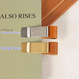 May include: Two pen clips attached to a book cover. The top clip is silver-toned stainless steel with an orange leather loop. The bottom clip is gold-toned brass with an orange leather loop. Text on the book cover reads ALSO RISES, Orange-Stainless, and Orange-Brass.