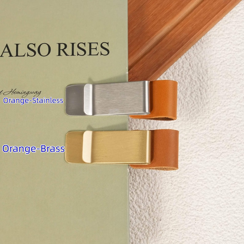 May include: Two pen clips attached to a book cover. The top clip is silver-toned stainless steel with an orange leather loop. The bottom clip is gold-toned brass with an orange leather loop. Text on the book cover reads ALSO RISES, Orange-Stainless, and Orange-Brass.