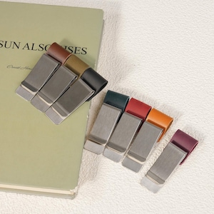 May include: A collection of metal money clips featuring leather accents in various colors including brown, tan, black, dark green, red, orange, and purple. Each clip has a brushed silver-tone finish and a rectangular shape for holding currency.