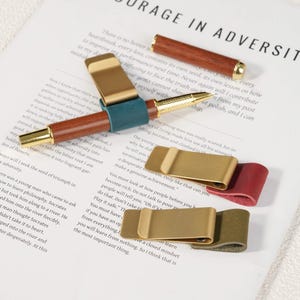 May include: A wooden pen with gold-tone accents and a matching cap, accompanied by three metal pen clips featuring teal, red, and olive green leather loops. The items are arranged on a white paper surface with printed text visible in the background.