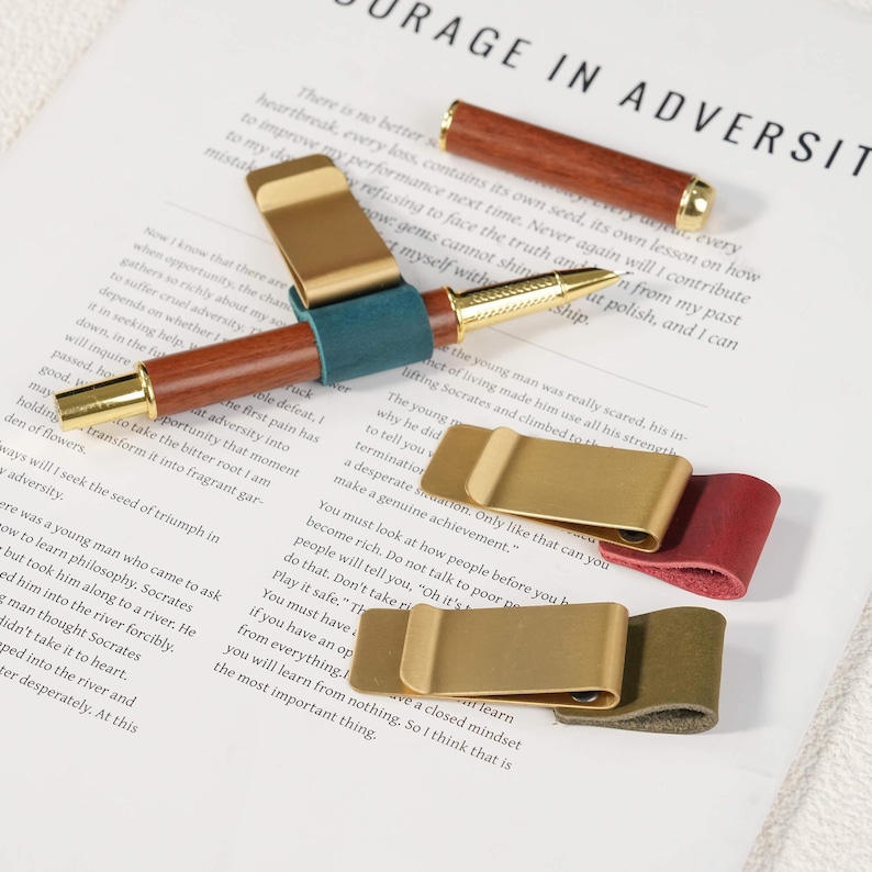 May include: A wooden pen with gold-tone accents and a matching cap, accompanied by three metal pen clips featuring teal, red, and olive green leather loops. The items are arranged on a white paper surface with printed text visible in the background.