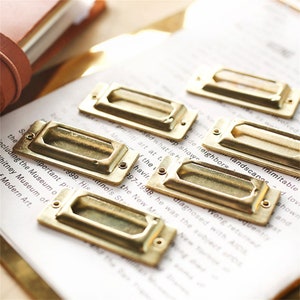 May include: Six gold-toned metal drawer pulls with a rectangular shape and a cut-out handle. The pulls are arranged in a staggered pattern on a white surface with text.