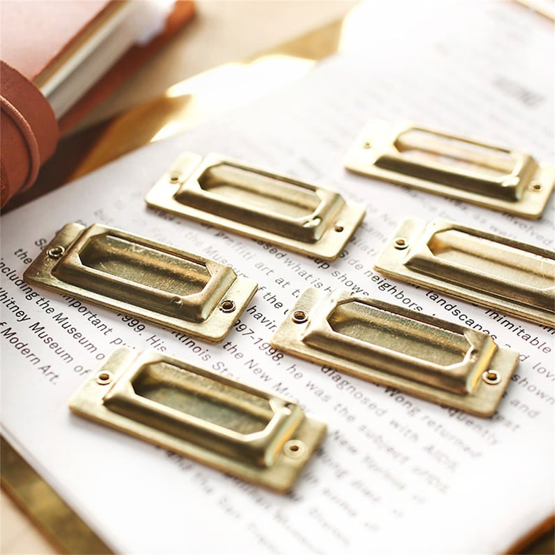 May include: Six gold-toned metal drawer pulls with a rectangular shape and a cut-out handle. The pulls are arranged in a staggered pattern on a white surface with text.