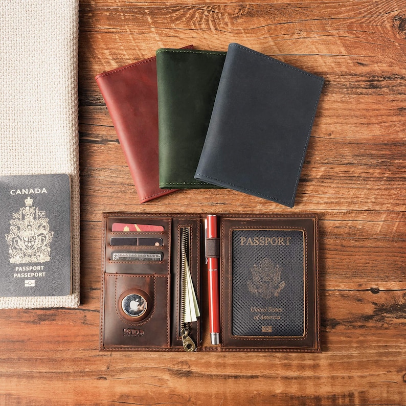 May include: Three leather passport covers in red, green, and navy blue are arranged above an open brown leather passport wallet. The wallet features card slots, a zippered pocket, a pen loop with a red pen, and a clear window holding a passport.