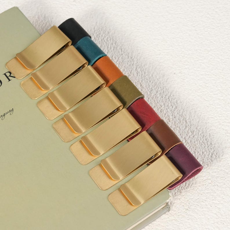 May include: A collection of eight metal money clips, each featuring a brushed gold finish and a leather strap in a different color, including black, teal, orange, olive green, red, brown, and purple, attached to the edge of a book.