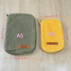 May include: Two zippered pouches, one olive green (16" x 9.84") and one yellow (4.72" x 8.26"). The green pouch is labeled "A5" and the yellow pouch is labeled "A6". Both have a small leather tag with a name.
