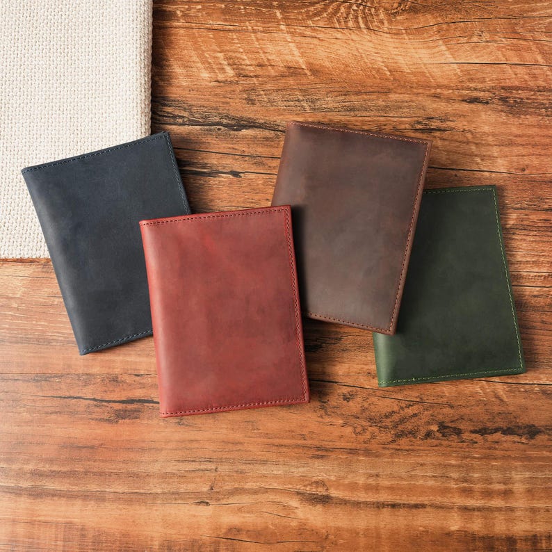 May include: Four rectangular leather passport holders or notebook covers in navy blue, dark red, brown, and forest green. Each item features a smooth, matte finish with visible stitching along the edges, arranged in a cluster on a wooden surface.