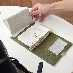 May include: An open, olive green planner with gold-coloured rings and a clear pocket. The planner's pages are white with the text "LIFE IS WHAT YOU MAKE IT". A hand is turning the pages of the planner.