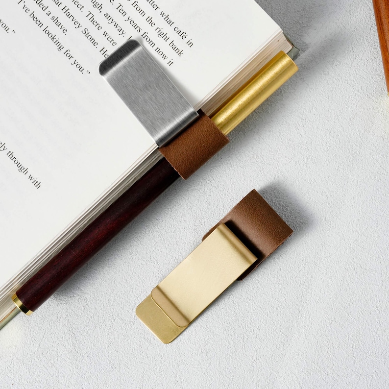 May include: Two metal and leather book page holders. One is silver with a brown leather accent, and the other is gold with a brown leather accent. The holders are on a book.