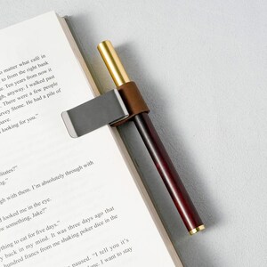 May include: A pen-shaped bookmark with a dark wood body, gold accents, and a silver clip. The bookmark is attached to the pages of a book. The pen-shaped bookmark is a unique accessory for book lovers.