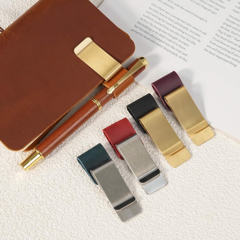 May include: A collection of five pen holders, each featuring a leather loop attached to a metal clip. One holder is attached to a brown leather notebook with a brown pen inserted. The other four clips are in dark green, red, black, and purple leather.