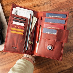 May include: A red leather travel wallet, open to display multiple compartments for cards, passports, and a pen. The wallet has a snap closure and an RFID-blocking feature. The interior includes a zippered pocket and multiple card slots.